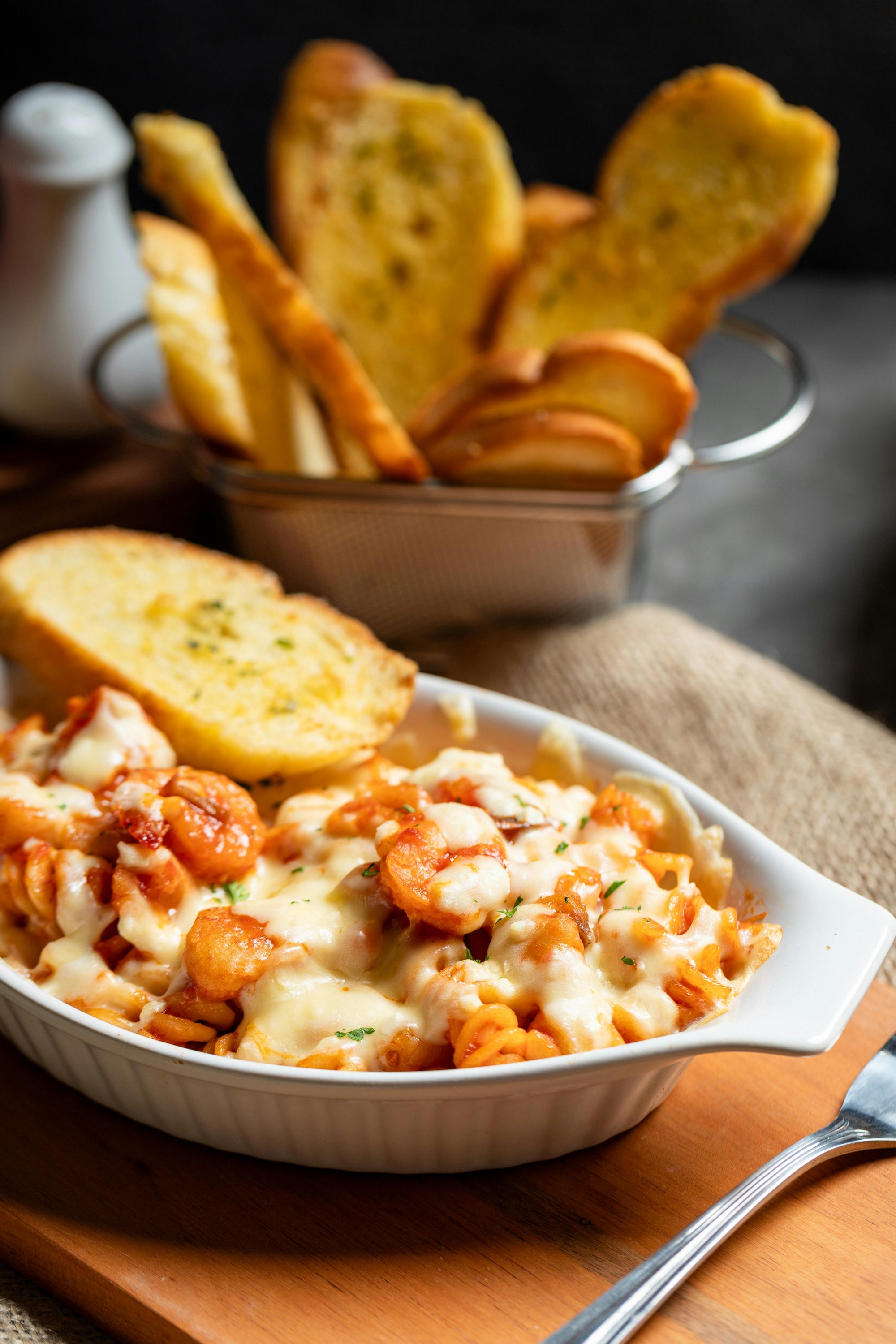 "Ofengebackene Pasta mit geschmolzenem Käse und Garnelen, serviert mit knusprigem Knoblauchbrot in einem weißen Auflaufform, auf einem Holzbrett angerichtet.